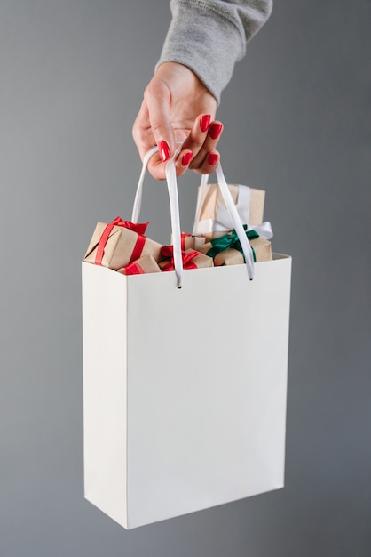 Foto beschnittenes bild der weiblichen hand mit den roten polierten nägeln, die einfache weiße einkaufstasche voller weihnachtsgeschenkboxen halten