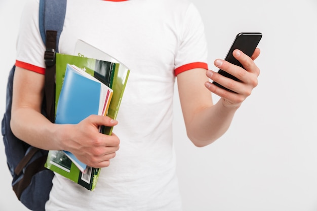 Beschnittenes Bild des europäischen Studentenjungen mit Rucksack, der viele bunte Übungsbücher hält und Handy lokalisiert auf Weiß hält
