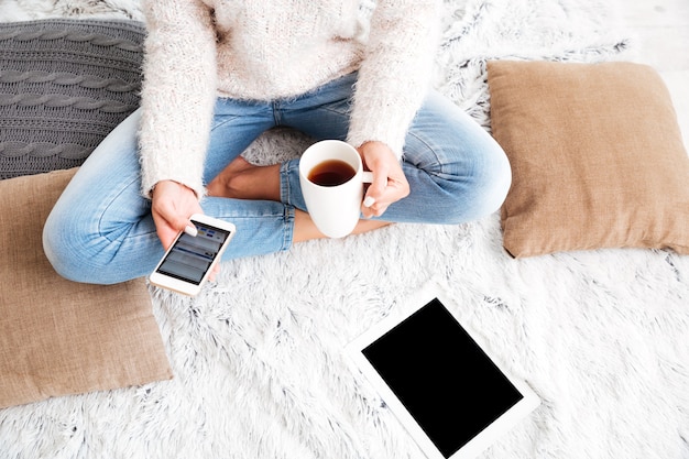 Beschnittenes Bild einer Frau, die Teetasse und leere Bildschirmtablette beim Sitzen auf dem Teppich zu Hause hält