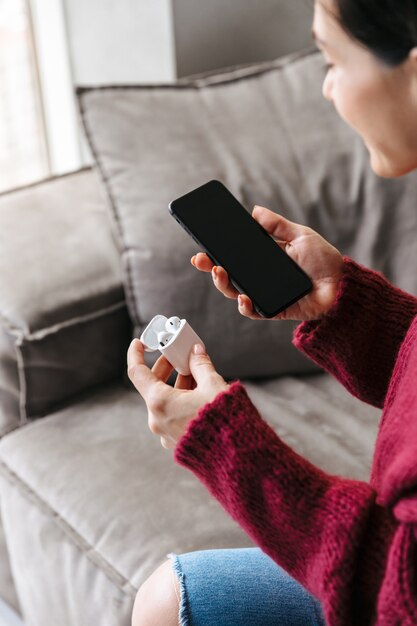 Beschnittenes Bild einer Frau drinnen im Haus auf Sofa, das Fall mit Kopfhörern und Handy hält.