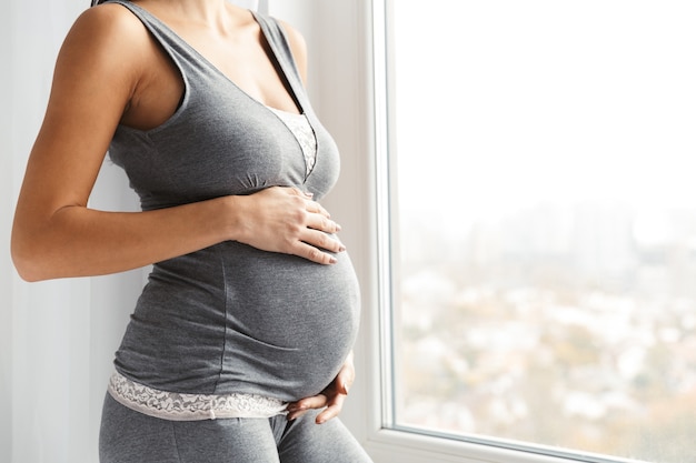 Beschnittenes Bild einer jungen schwangeren Frau, die am Fenster steht und Bauch berührt