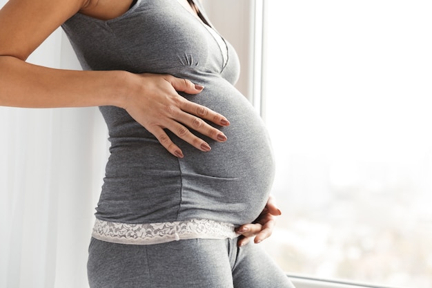 Beschnittenes Bild einer jungen schwangeren Frau, die am Fenster steht und Bauch berührt