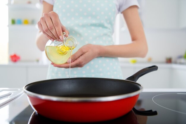 Beschnittenes Nahaufnahmefoto von Hausfrauenhänden, die Olivenölflasche halten, die warme Bratpfanne kocht, die leckere Abendessenvorbereitungsfamilientreffen-Weißlichtküche drinnen kocht