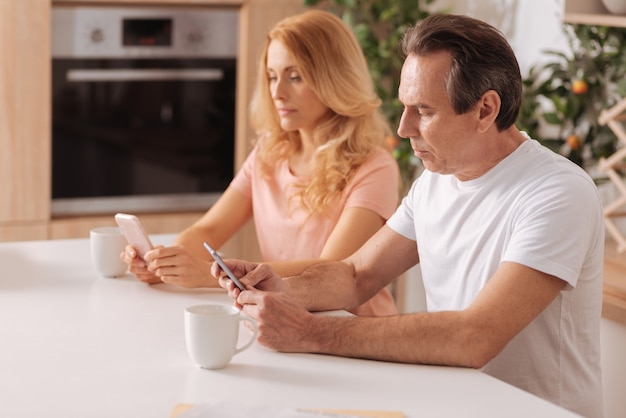 Besessenes modernes sorgloses Paar, das zu Hause sitzt und Smartphones benutzt, ohne aufeinander zu achten