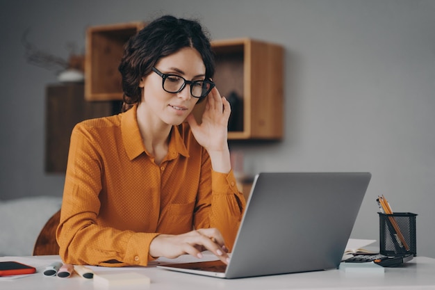 Besetzte Spanierin mit Brille sitzt am Schreibtisch und tippt während der Fernarbeit zu Hause auf dem Laptop