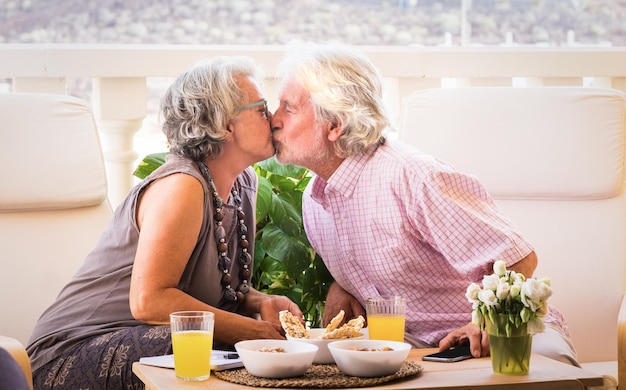 Un beso de amor entre dos personas mayores pensionistas con cabello blanco y gris Pareja sentada al aire libre en la terraza Un momento de relajación con comida y bebida Luz brillante