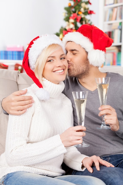 Beso de Navidad. Hermosa joven sosteniendo una copa de champán y sonriendo mientras su novio besa su mejilla