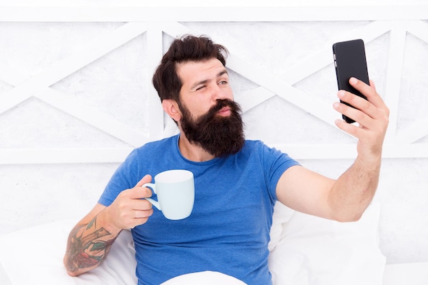 Beso de selfie. Hombre barbudo tomar selfie en la cama. Sonrisa inconformista a cámara selfie en teléfono móvil. Disfrutando de la sesión de selfies. Rutina del café matutino. Vida moderna. Nueva tecnología. Red social.