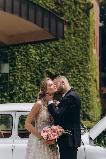 Beso sensual de los dos en el día de su boda cerca de un auto retro con flores.