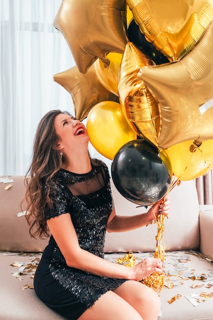 Besonderer Tag. Amüsiertes brünettes Mädchen, das auf Sofa im schwarzen Pailletten-Minikleid sitzt, lacht und Bündel goldener Luftballons hält.