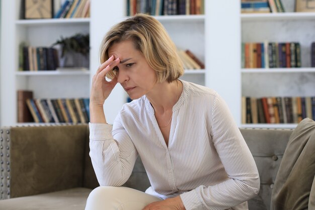 Besorgte Frau mittleren Alters, die auf einem Sofa im Wohnzimmer sitzt.