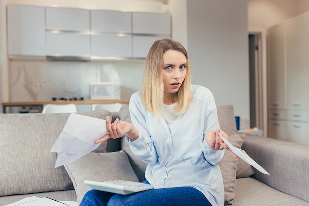 Besorgte Hausfrau zahlt überhöhte Rechnungen, während sie zu Hause auf dem Sofa sitzt, frustriert über die Arbeit und das monatliche Einkommen