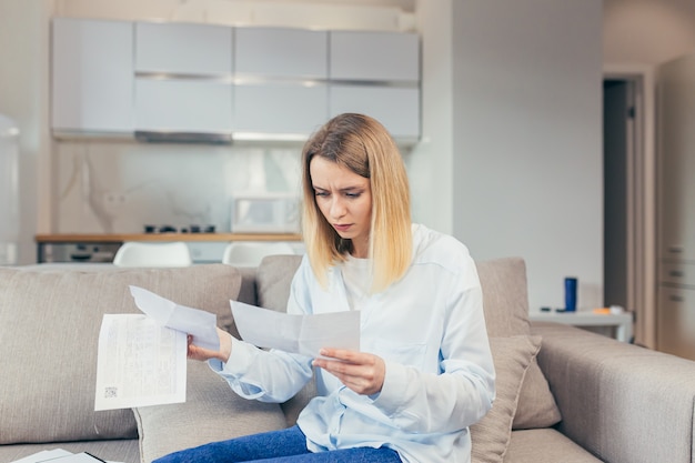 Besorgte Hausfrau zahlt überhöhte Rechnungen, während sie zu Hause auf dem Sofa sitzt, frustriert über die Arbeit und das monatliche Einkommen