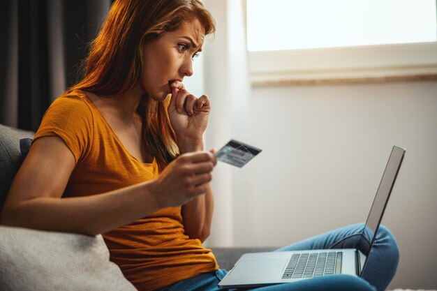 Besorgte junge Frau beim Online-Shopping auf ihrem Laptop, während sie zu Hause auf dem Sofa sitzt.