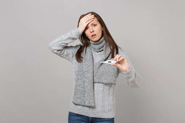 Besorgte junge Frau in grauem Pullover, Schal Hand auf die Stirn legen, Thermometer auf grauem Hintergrund isoliert halten. Gesunder Lebensstil, Behandlung kranker Krankheiten, Konzept der kalten Jahreszeit. Kopieren Sie Platz.