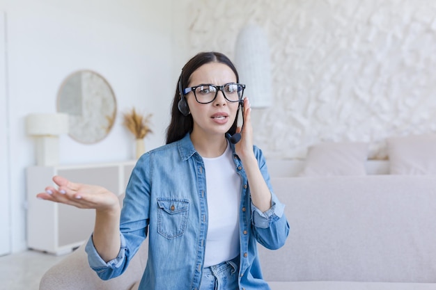 Besorgte junge Frau sitzt zu Hause auf dem Sofa im Headset und spricht online über Mikrofon