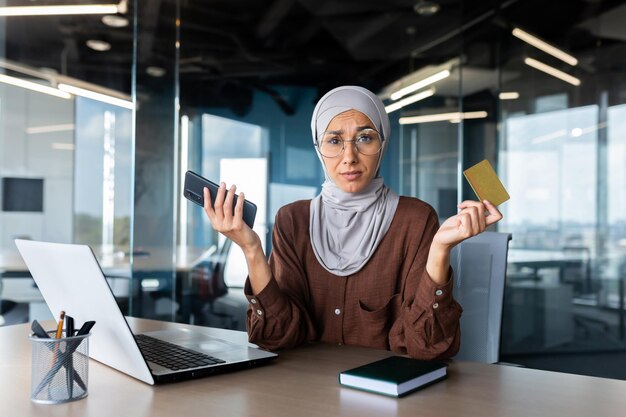 Besorgte und verärgerte junge muslimische Geschäftsfrau im Hijab, die Telefon und Kreditkarte in der Hand hält