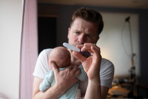 Besorgter junger Vater, der auf das Thermometer blickt und sein neugeborenes Baby in den Händen hält.