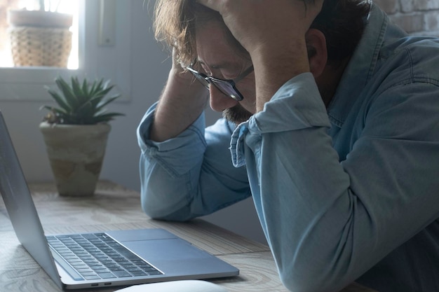 Besorgter und verzweifelter gestresster Arbeiter am Schreibtisch, der sich mit erschöpfter Miene den Kopf hält Seitenansicht eines Geschäftsmannes, eines Kleinunternehmens mit Problemen und Pleite vor einem Laptop-Computer online