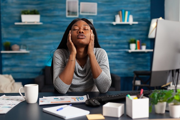 Besorgter unzufriedener afrikanischer Student, der unter Kopfschmerzen Migräne leidet, sitzt am Schreibtisch im Wohnzimmer ...