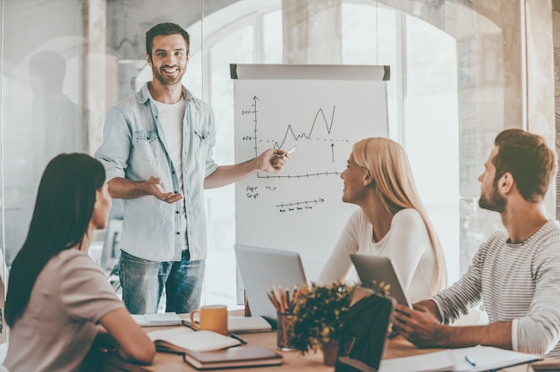 Besprechung des Unternehmensfortschritts. Selbstbewusster junger Mann, der in der Nähe des Whiteboards steht und auf ein Diagramm zeigt, während seine Kollegen am Schreibtisch sitzen