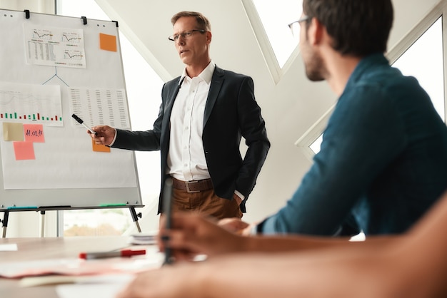 Besprechung von Geschäftsideen. Reifer Geschäftsmann, der nahe Whiteboard steht und seinen jungen Kollegen etwas erklärt. Zusammenarbeit. Treffen