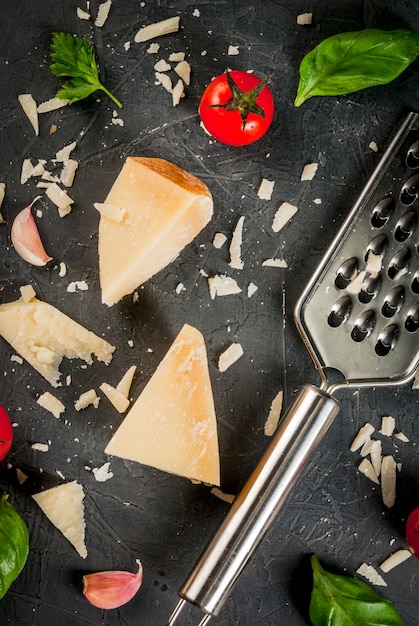 Bestandteile der italienischen Küche Parmesankäse zerrieben und ein Stück, mit einer Reibe, Basilikumblättern, Knoblauch und Kirschtomaten auf einer dunklen konkreten Tabelle. Ansicht von oben