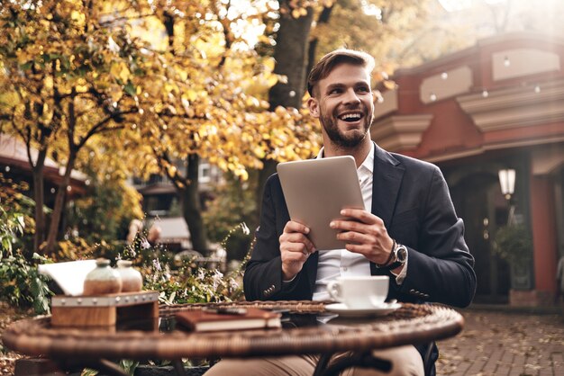 Beste Ergebnisse erzielen. Gut aussehender junger Mann in eleganter Freizeitkleidung mit digitalem Tablet und mit einem Lächeln wegschauen, während er im Restaurant im Freien sitzt