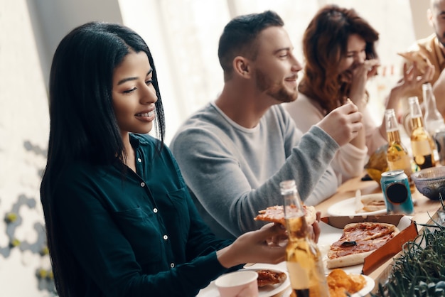 Beste Freunde. Gruppe junger Leute in Freizeitkleidung, die Pizza isst und lächelt, während sie eine Dinnerparty im Haus veranstalten