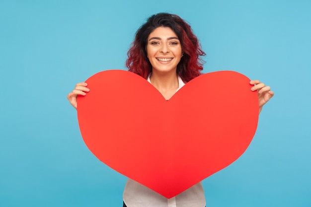 Beste Wünsche zum Valentinstag. Begeisterte hübsche hübsche Frau mit ausgefallenen roten Haaren, die ein großes Herz hält, Symbol der Liebe und Zuneigung, romantischer Urlaub. Studioaufnahme im Innenbereich isoliert auf blauem Hintergrund