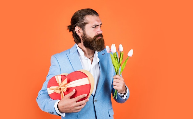 Bester bärtiger Mann zum Valentinstag in eleganter Jacke hält Blumen Hochzeitstag glücklicher Valentinstag Braut Bräutigam mit Blumenstrauß und Schachtel Hipster-Mann hält Tulpenblumen Liebesgeschenk für ein romantisches Date