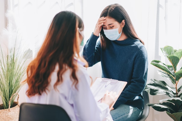 Besuch eines asiatischen Arztes und Untersuchung einer jungen erwachsenen Frau im Krankenhaus mit Kopfschmerzen Der Arzt untersucht und konsultiert die Gesundheitsversorgung mit einer Maske zum Schutz der Covid19-Pandemie