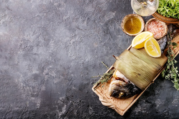 Besugo o dorado pescado cocido fresco eviscerado a la parrilla servido con limón, ensalada verde, romero, aceite de oliva y sal rosa en un plato de cerámica envuelto en hojas de bambú sobre una superficie de textura oscura. Endecha plana