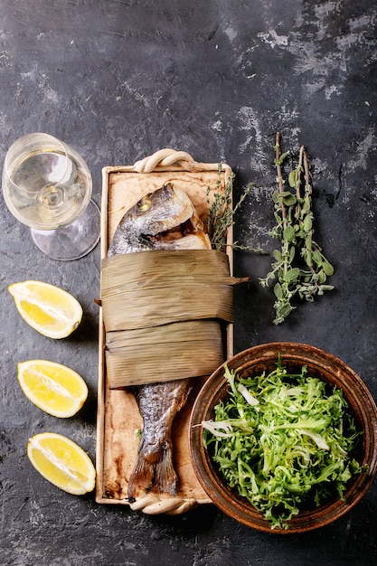 Foto besugo o dorado pescado fresco cocido a la parrilla, servido con limón, ensalada verde, romero, aceite de oliva, vino blanco y sal rosa en un plato de cerámica envuelto en hojas de bambú sobre una superficie oscura.