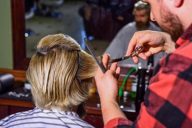 Betrachten Sie sich gewarnt Hipster-Kunde bekommt Haarschnitt Friseurschere Kerl mit lang gefärbten blonden Haaren Nahaufnahme Rückansicht Haare schneiden Friseur macht Frisur für bärtigen Mann Barbershop-Hintergrund