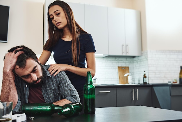 Betrunkener Ehemann. Dunkelhaarige süchtige Frau, die Verzweiflung fühlt, während sie in der Nähe ihres betrunkenen Mannes steht. Er sitzt in der Küche, erschöpft. Auf dem Tisch neben ihm stehen leere Flaschen.