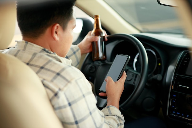 Betrunkener Mann, der eine Flasche Bier hält und ein Smartphone benutzt, während er ein Auto fährt, das sich für betrunkenes Fahren einsetzt, um Unfälle auf der Straße zu vermeiden. Konzept nicht trinken und fahren