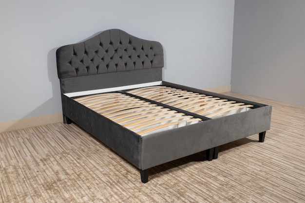 Bett mit grauer Stoffpolsterung und Holzrahmen ohne Matratze in einem leeren Raum