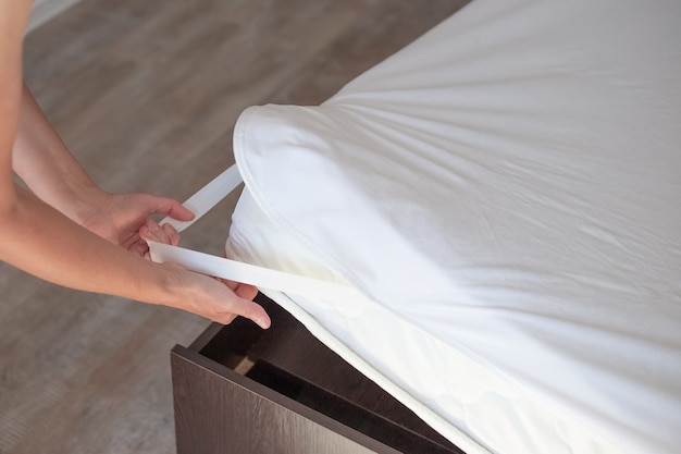 Bett mit hoher Matratze. Die Frau zieht einen schützenden wasserabweisenden Matratzenbezug über.