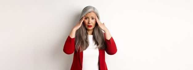 Beunruhigte asiatische Unternehmerin, die Kopfschmerzen hat, die den Kopf berühren, und die Stirn runzelt, wenn sie über weißem Ba steht