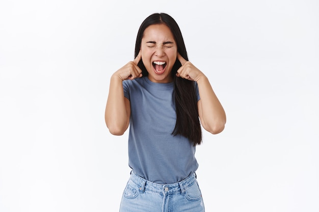 Beunruhigtes und sattes asiatisches Teenager-Mädchen mit langen dunklen Haaren, das schreiend die Beherrschung verliert, Emotionen nicht kontrollieren kann, Augen schließen und schreien, enge Ohren mit Fingern können die Leute nicht ertragen, die um sie herum kämpfen