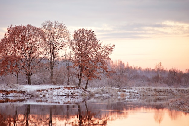 Bewölkte Herbstdämmerung. Erster Schnee auf dem Herbstfluss. Eichen am Flussufer.