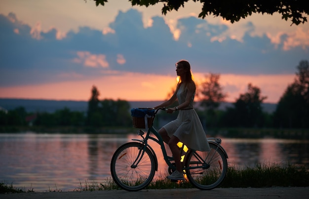 Bezaubernde Sonnenuntergangslandschaft Silhouette einer Frau im Kleid fährt ein Retro-Fahrrad mit Korb auf der Straße in der Nähe des Flusses