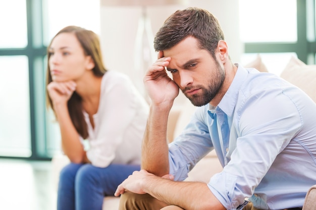 Beziehungszusammenbruch. Depressiver junger Mann, der die Hand am Kopf hält und wegschaut, während die Frau hinter ihm auf der Couch sitzt