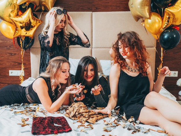 BFF-Treffpunkt. Urban Girls Freizeit und Lifestyle. Glitzer Konfetti und Luftballons Dekor. Junge Frauen in Schwarz, die auf Bett entspannen.