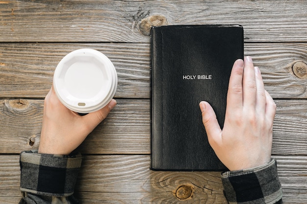 Foto biblia y una taza de café de papel en manos masculinas en una vista superior de fondo de madera