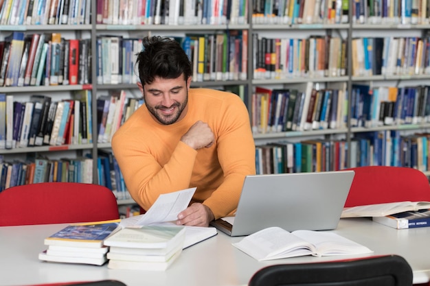 En la biblioteca Apuesto estudiante masculino con computadora portátil y libros que trabajan en una biblioteca universitaria de secundaria con poca profundidad de campo