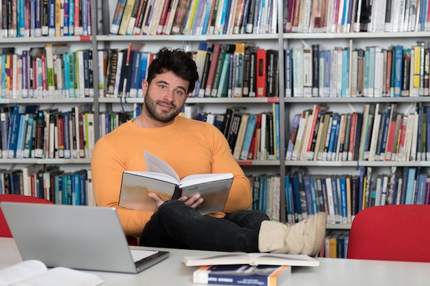 En la biblioteca Apuesto estudiante masculino con computadora portátil y libros que trabajan en una biblioteca universitaria de secundaria con poca profundidad de campo