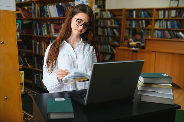 Biblioteca da universidade Linda garota caucasiana inteligente usa laptop escreve notas para estudo de redação de papel para tarefa de classe estudantes focados aprendendo estudando para exames universitários vista lateral retrato