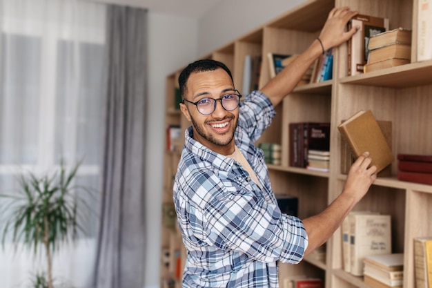 Biblioteca doméstica feliz homem árabe tirando livro da estante pesquisando e escolhendo manual sorrindo para